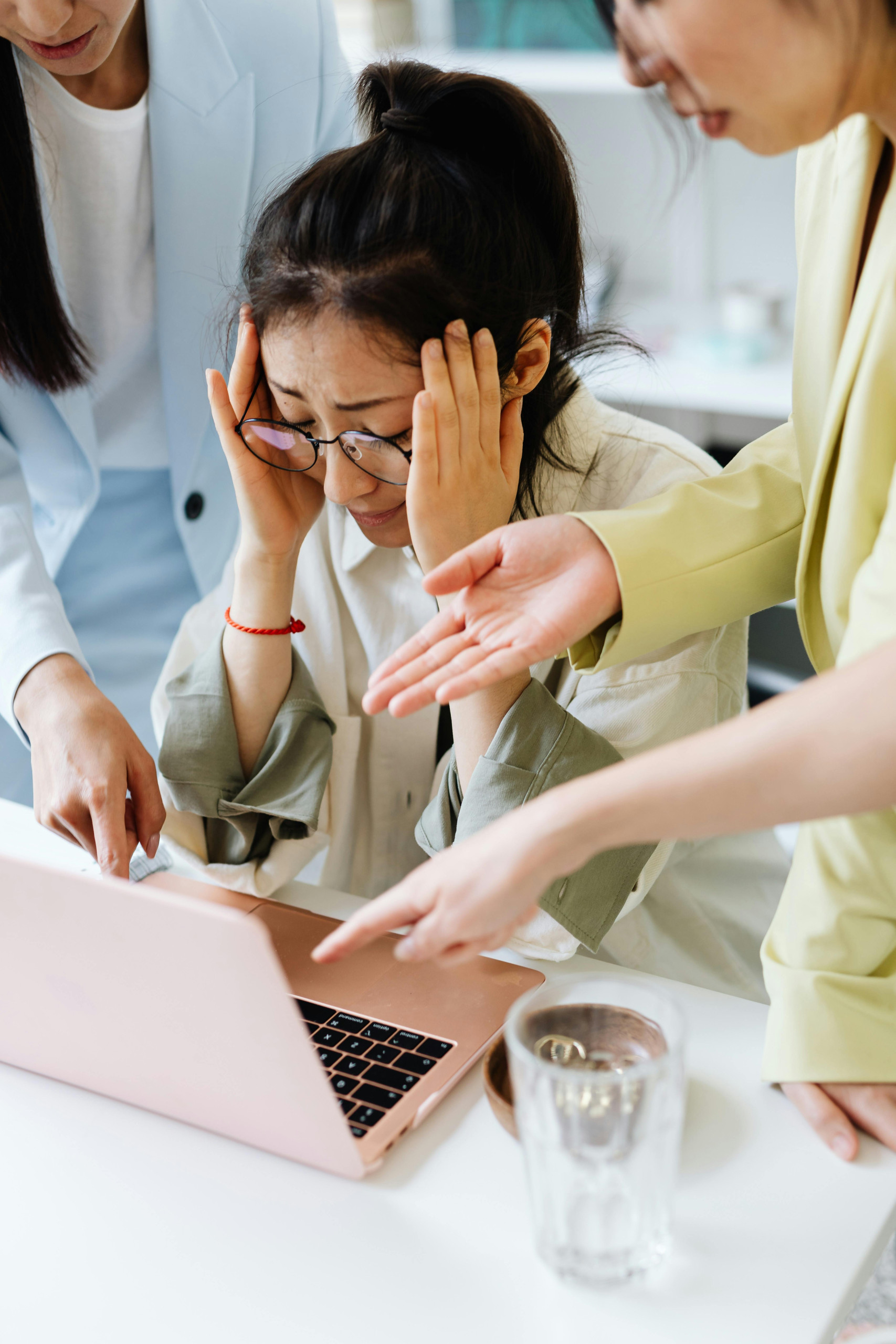 A stressed woman holding her head looking at her laptop while colleagues point at the screen